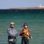 Two people kiteboarding with safety gear on a sunny day at sea, wearing helmets and wetsuits.