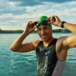 Confident male athlete adjusting goggles at a lake during a sunny day, ready for training.